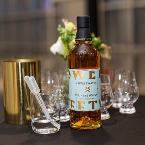 Bottle of Sweetwater Waiheke Whisky on a table with glasses and a gold container.