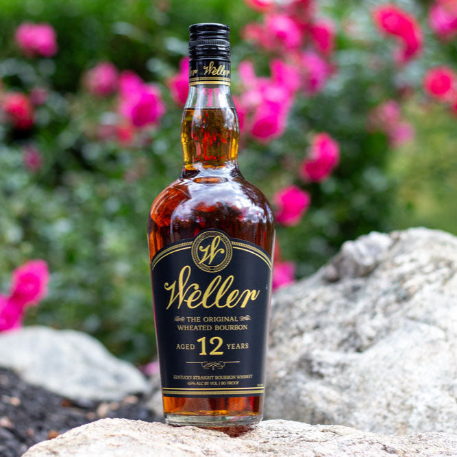 Weller Whiskey bottle standing on a rock in front of a bush of flowers