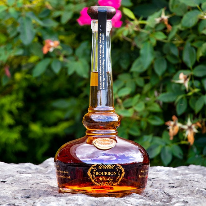 Load image into Gallery viewer, A bottle of Willett Pot Still Reserve Bourbon Whiskey standing on some rocks with a flowery bush as the background