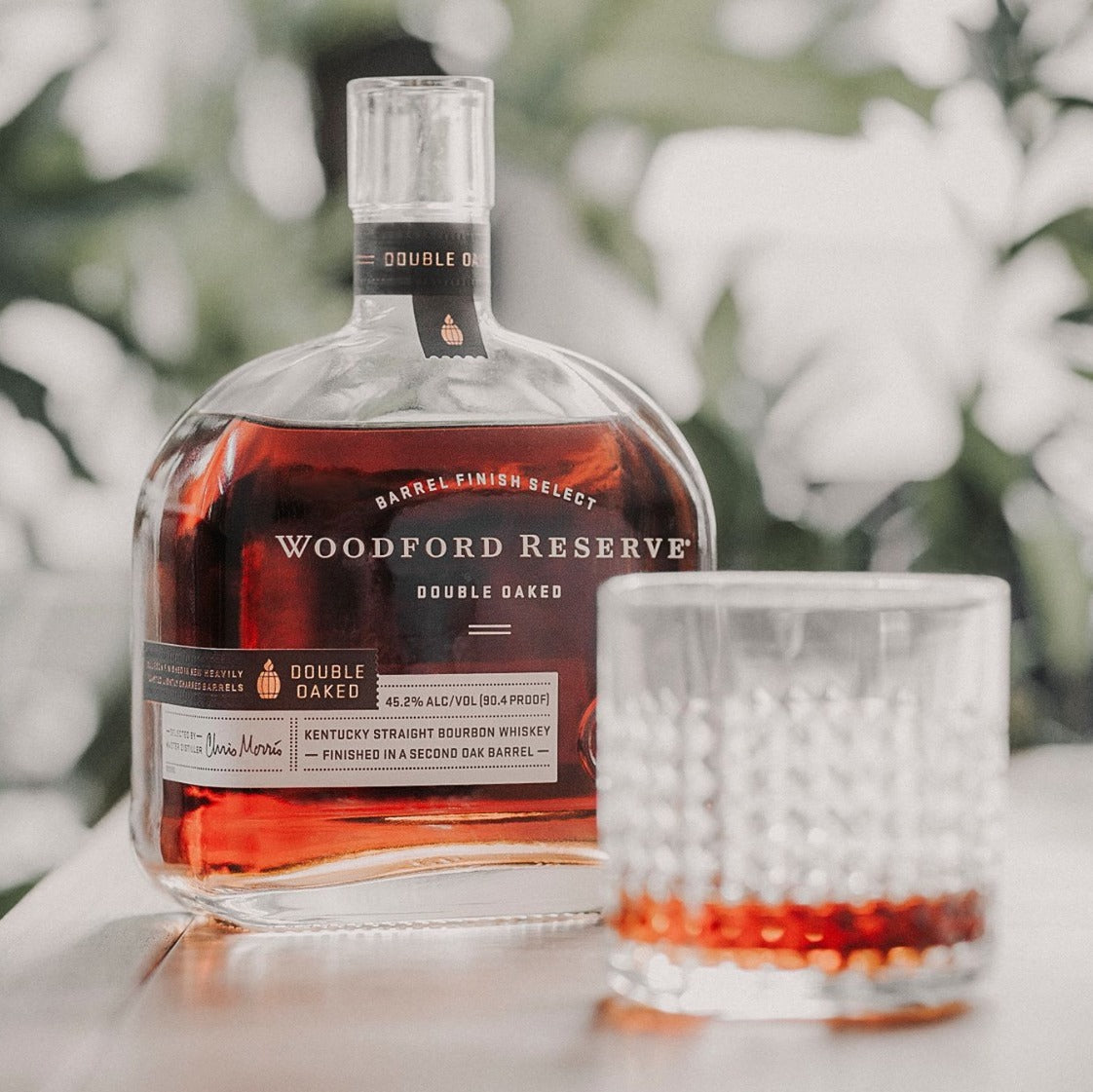 Load image into Gallery viewer, A bottle of Woodford Reserve Double Oaked Bourbon on a white table with a tumbler with a green blurry background