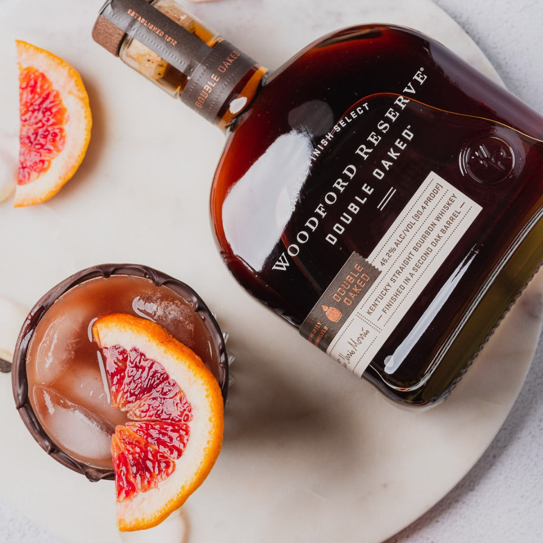 Load image into Gallery viewer, A bottle of Woodford Reserve Double Oaked Bourbon laying down beside a whiskey cocktail with a blood orange wedge