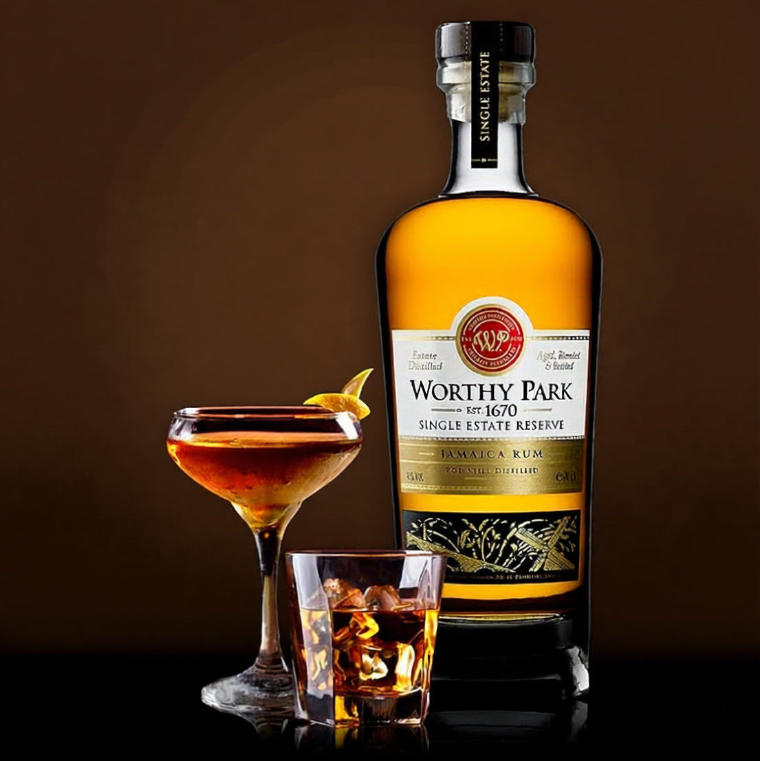 Load image into Gallery viewer, A bottle of Worthy Park Single Estate Reserve Rum stands on a dark reflective surface beside a filled coupe glass and an rocks tumbler with ice, all showing the amber liquor. The background is a dark brown studio backdrop.