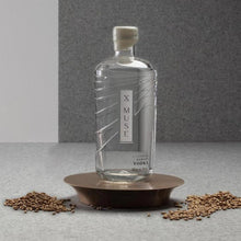 Load image into Gallery viewer, Bottle of X Muse Vodka on a gray surface with barley grains around it.