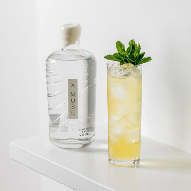 Bottle of X Muse vodka next to a cocktail with mint leaves on a white surface.
