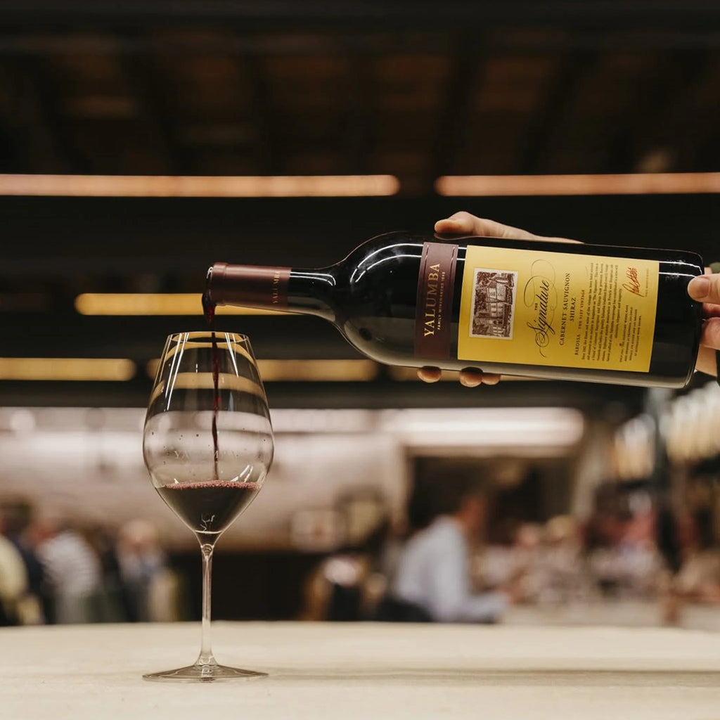 Person pouring a bottle of Yalumba The Signature Cabernet Sauvignon Shiraz from a bottle into a glass with a blurred bar background