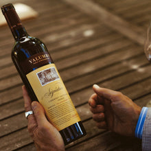 Load image into Gallery viewer, Person holding a bottle of Yalumba The Signature Cabernet Sauvignon Shiraz with a glass of wine in the background.