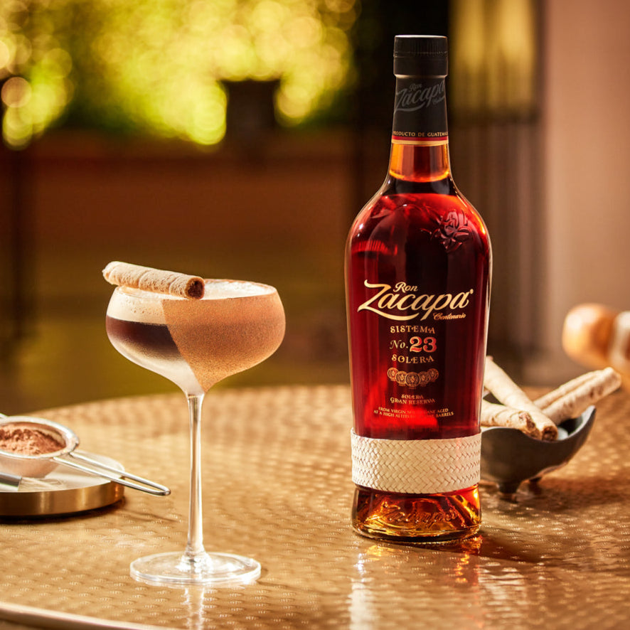 Bottle of Ron Zacapa 23 rum on a table with a cocktail and cinnamon sticks.