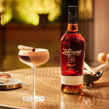 Load image into Gallery viewer, Bottle of Ron Zacapa 23 rum on a table with a cocktail and cinnamon sticks.
