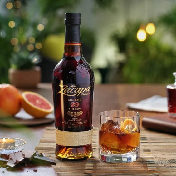 Bottle of Ron Zacapa 23 aged rum on a table with a glass of cocktail and fruit.