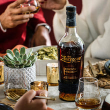 Load image into Gallery viewer, Bottle of Ron Zacapa Edicion Rum on a table with people and food in the background