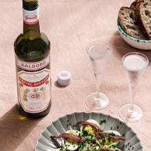 Load image into Gallery viewer, Deviled eggs with bacon on a plate, bottle of aalborg-taffel-akvavit, and two glasses on a pink surface.