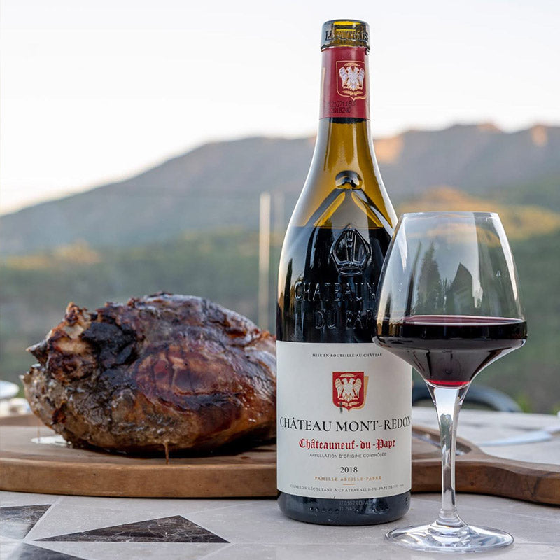 Load image into Gallery viewer, A bottle of Château Mont-Redon Châteauneuf-du-Pape red wine with a glass of red wine and a roasted ham on a cutting board, set against a blurred outdoor mountain background.