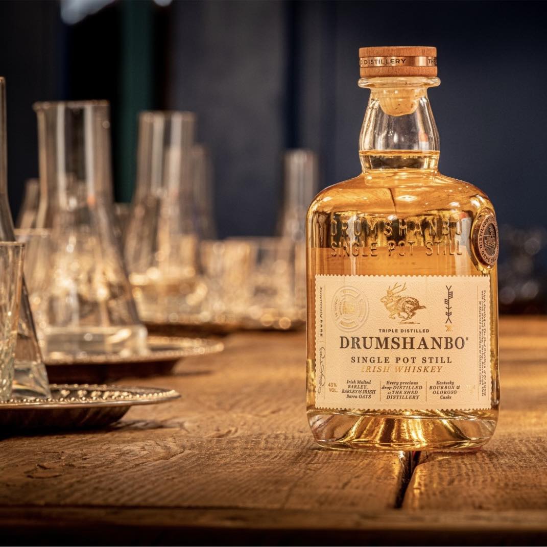 Load image into Gallery viewer, Bottle of Drumshanbo whiskey on a wooden table with glasses in the background