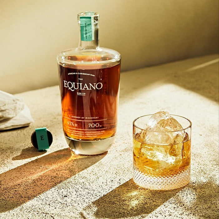 Load image into Gallery viewer, Bottle of Equiano rum and a glass with ice on a sandu wooden surface