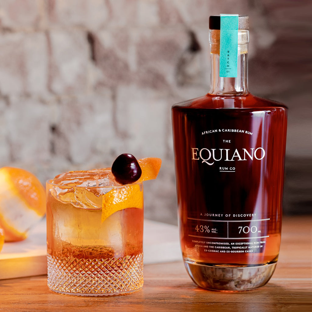 Bottle of Equiano rum with a cocktail garnished with an orange and cherry on a wooden surface.