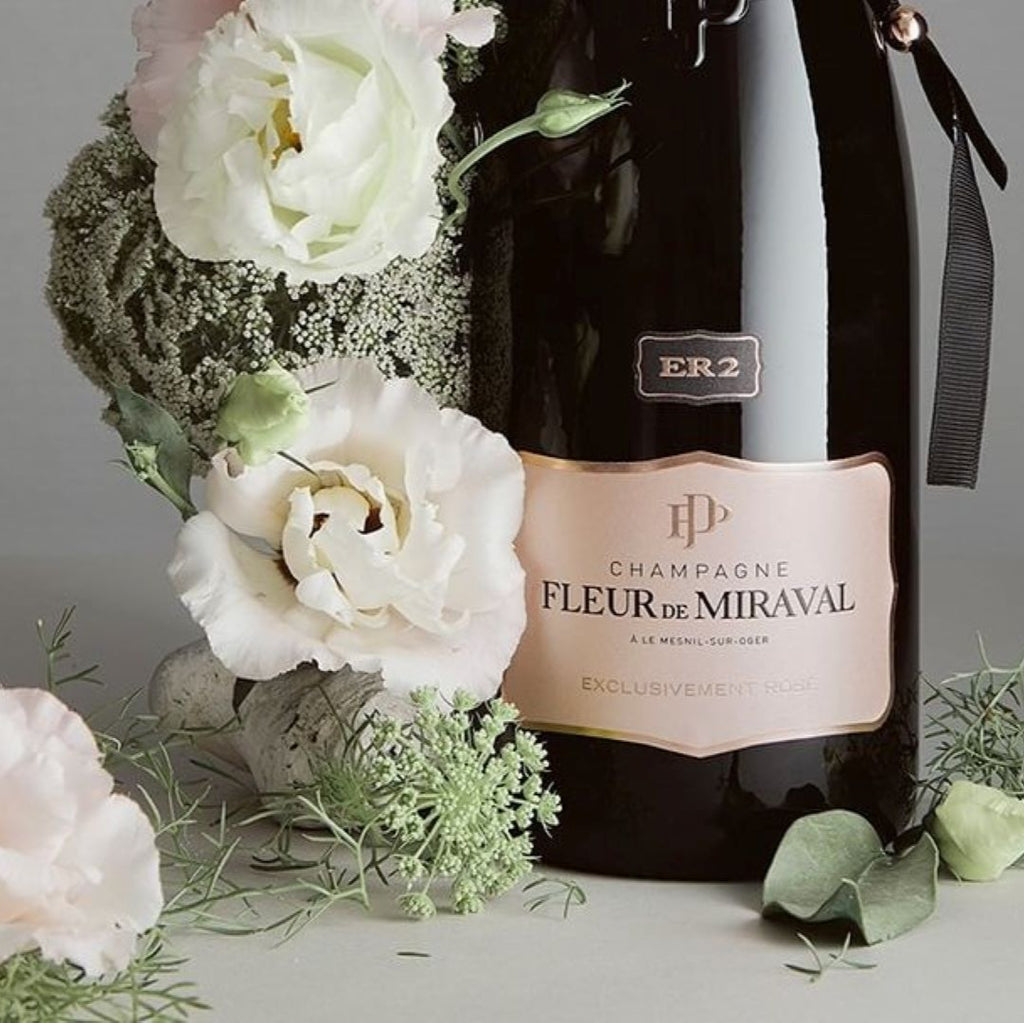 Champagne bottle with 'Fleur de Miraval' label surrounded by flowers on a grey background