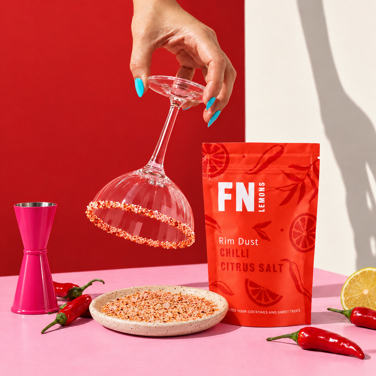 Load image into Gallery viewer, A hand with bright blue nail polish holds a clear wine glass rimmed with orange-red chili salt, next to a red stand-up pouch labeled 'FN Lemons Rim Dust Chilli Citrus Salt,' with red chili peppers and a pink jigger on a pink and red studio background.