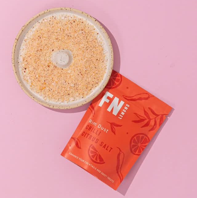 Load image into Gallery viewer, A flat-lay of a round ceramic dish filled with coarse sea salt next to a bright orange packet of FN Lemon's Rim Dust Chilli Salt on a solid pink background.