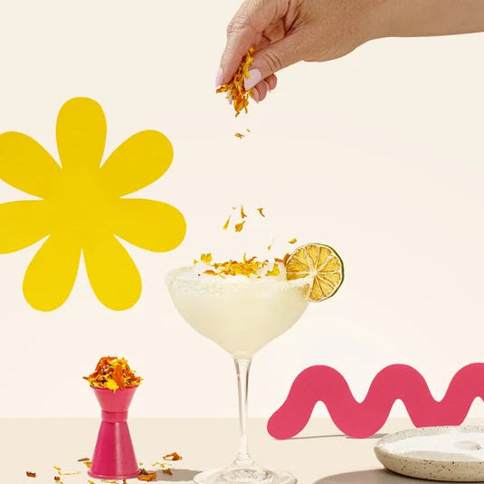 Load image into Gallery viewer, A hand sprinkles FN Lemon Summer Bloom dried flower florets onto a margarita in a coupe glass, with a lime wheel garnish on the rim. The scene includes decorative yellow and pink abstract shapes on a warm cream background.