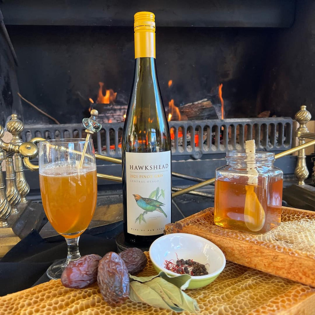 Wine bottle, glass of amber liquid, honeycomb, and honey dipper on a table with a fireplace in the background.