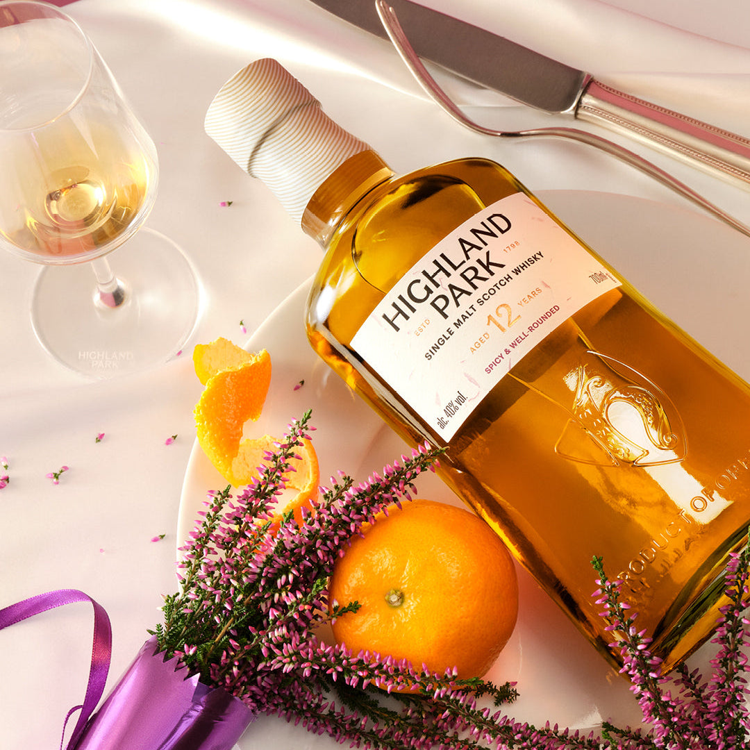Load image into Gallery viewer, Highland Park 12 year old whisky bottle with a glass, orange, and flowers on a white surface