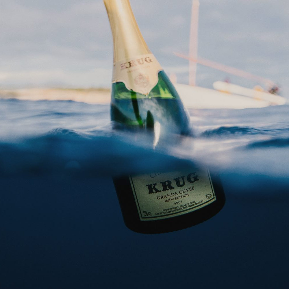 Krug champagne bottle partially submerged in water with a sailboat in the background
