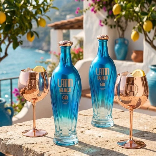 Load image into Gallery viewer, Two blue Latin Beach Gin bottles and two rose-gold stemmed gin goblets with lemon garnishes, arranged on a stone table with a Mediterranean seaside background.
