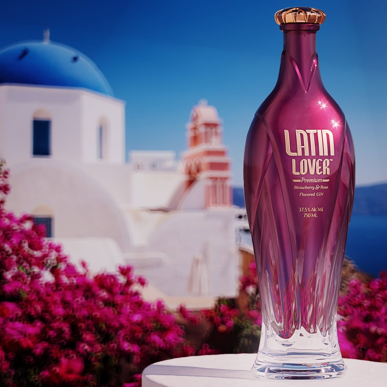 Load image into Gallery viewer, A faceted purple glass bottle of Latin Lover pink Gin with a gold cap, set against a blurred background of Santorini with a blue-domed church, white buildings, and bright pink bougainvillea flowers.