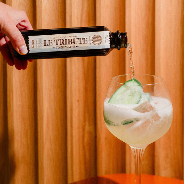 Load image into Gallery viewer, Bottle of 'Le Tribute' tonic water being poured into a glass with a drink and cucumber slice against a wooden background