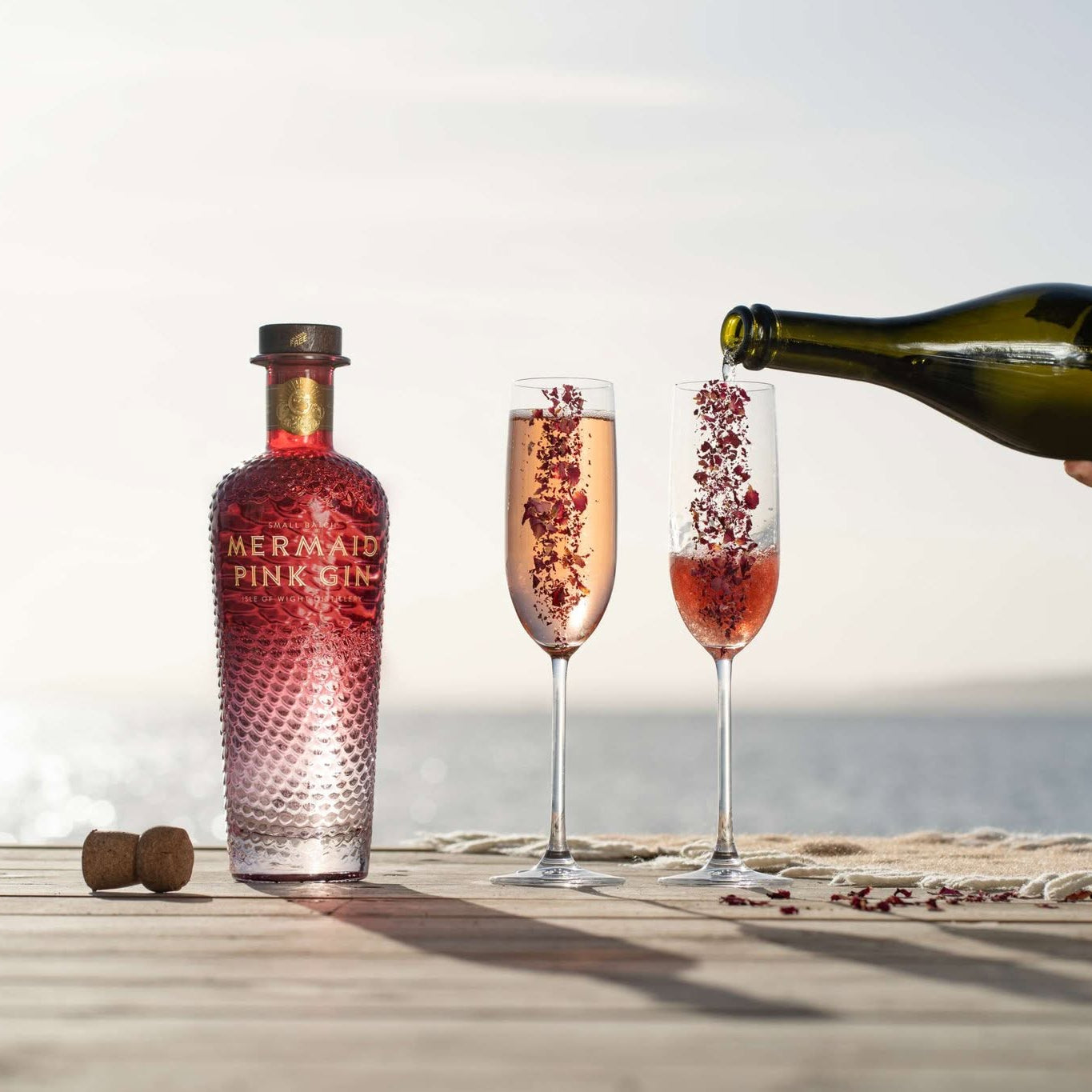 Load image into Gallery viewer, A bottle of Mermaid Pink Gin stands on a wooden deck beside two filled champagne flutes, with a hand pouring sparkling pink drink from a green bottle into one glass, against a bright seaside background.