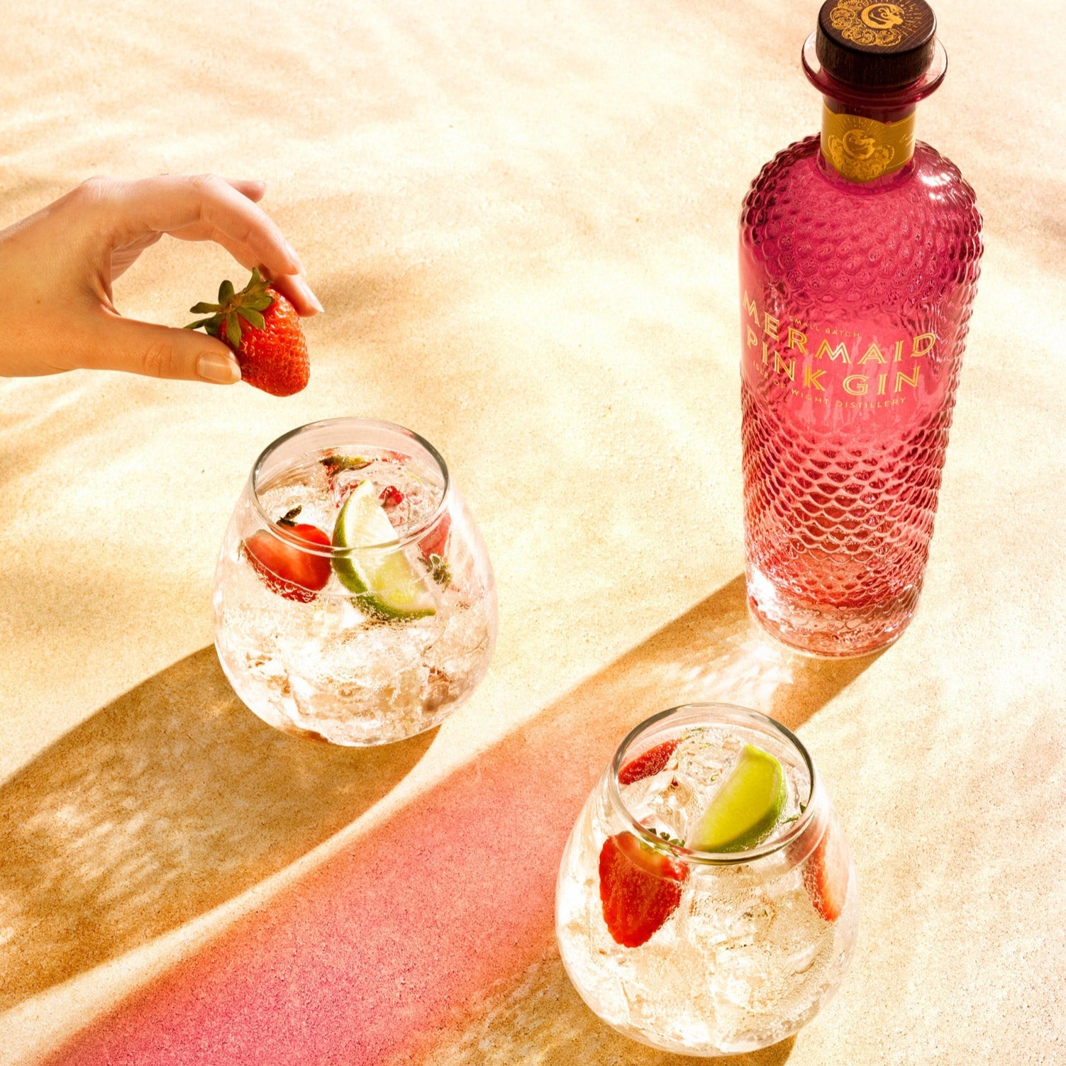 Load image into Gallery viewer, Bottle of Mermaid Pink Gin with two glasses filled with a pink drink, strawberries, and lime on a textured surface.