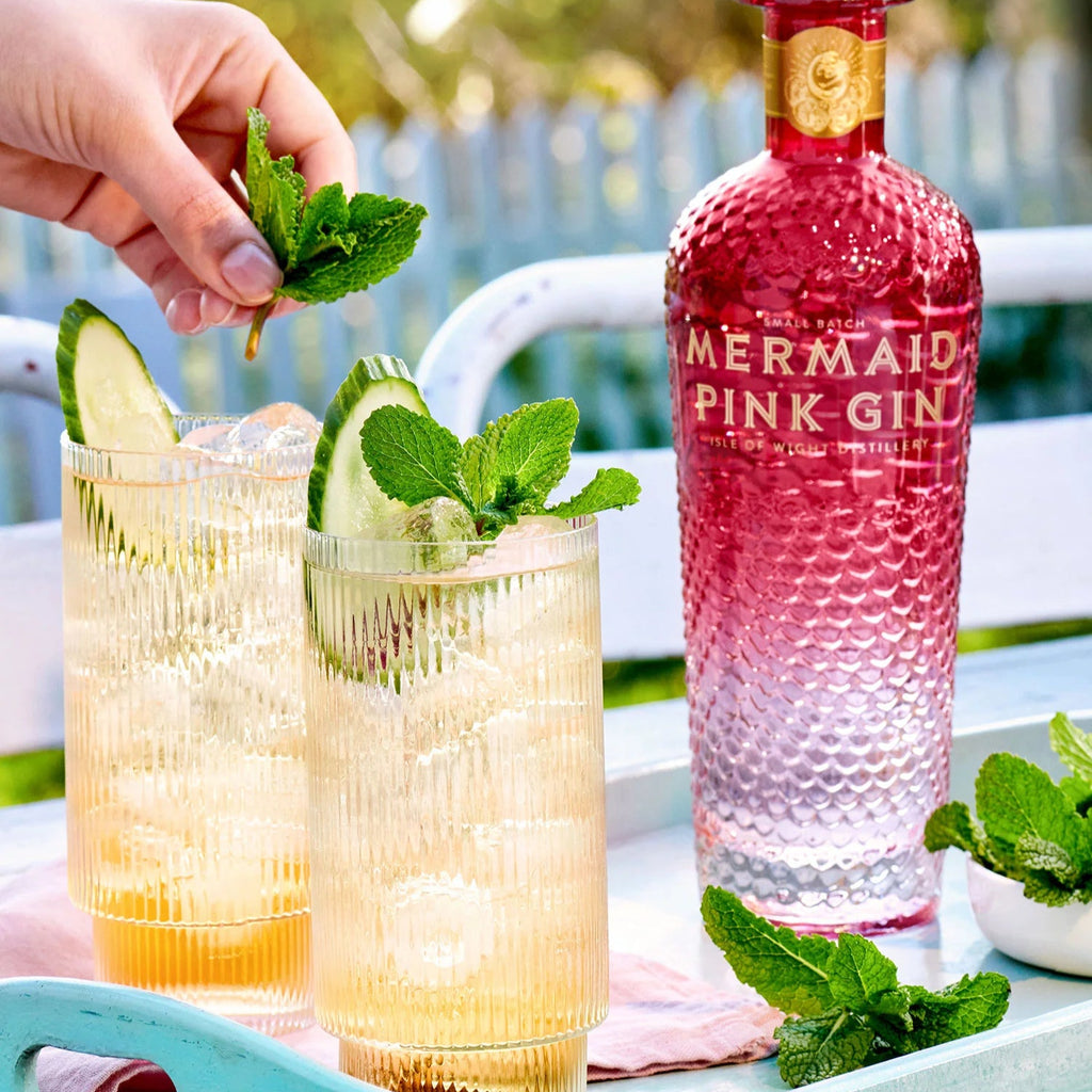 Two glasses of pink Mermaid gin cocktails with garnishes on a tray, accompanied by a bottle of Mermaid Pink Gin.