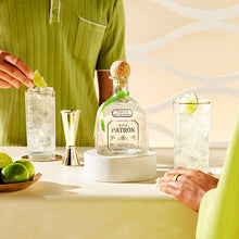 Load image into Gallery viewer, Silver Patron tequila bottle with a glass of lime garnished cocktail on a table.