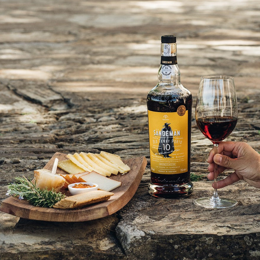 A hand holding a glass of port and a bottle of port and a cheeseboard on a stone stairway