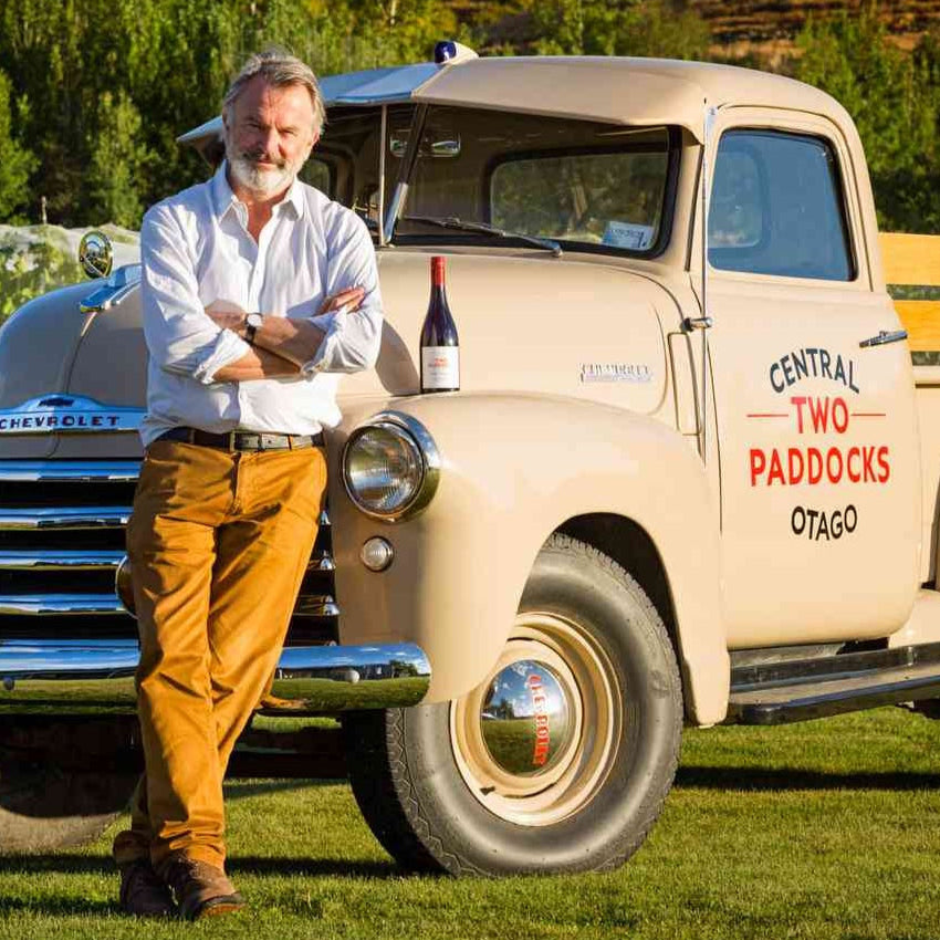 Actor Sam Neill standing next to a vintage truck with a bottle of Central Otago Two Paddocks Pinot Noir in a scenic outdoor setting.