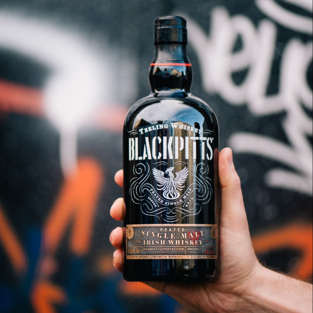 Load image into Gallery viewer, Bottle of Teeling Blackpitts Irish whiskey held in a hand with a graffiti-style background