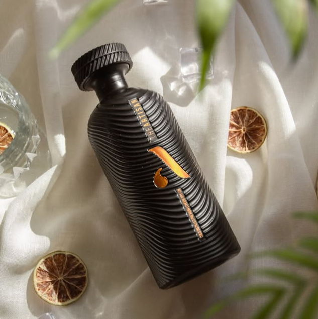 Black bottle of Artisan dry gin with orange label on a white fabric background with dried oranges and leaves.