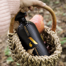Load image into Gallery viewer, Black bottle of artisan gin with a yellow label in a woven basket outdoors