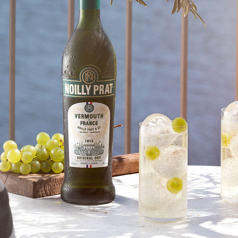 A bottle of Noilly Prat Original Dry Vermouth and some cocktails on a table with a blue background