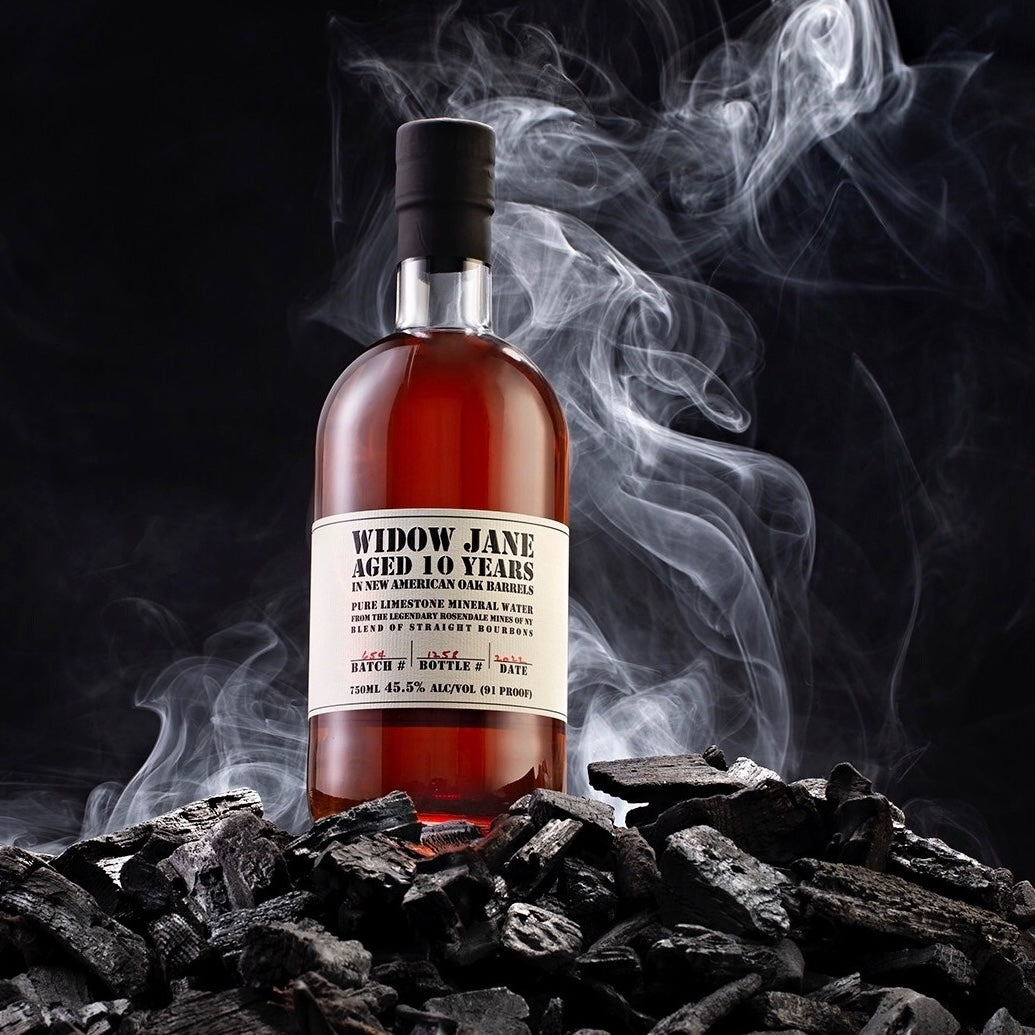 Load image into Gallery viewer, A bottle of Widow Jane 10YO Straight Bourbon standing on charcoal with a smoky background 