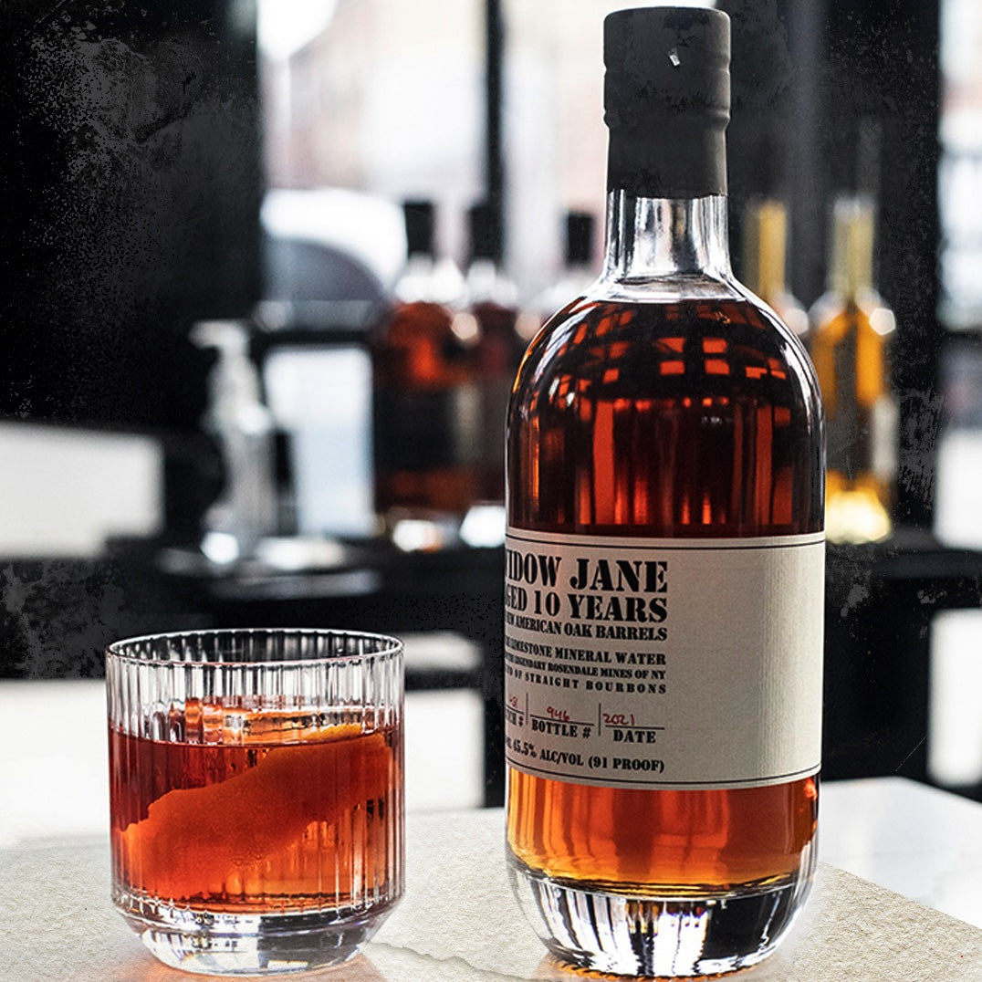 Load image into Gallery viewer, A bottle of Widow Jane 10YO Straight Bourbon on a table together with a tumbler full of whiskey with a black background